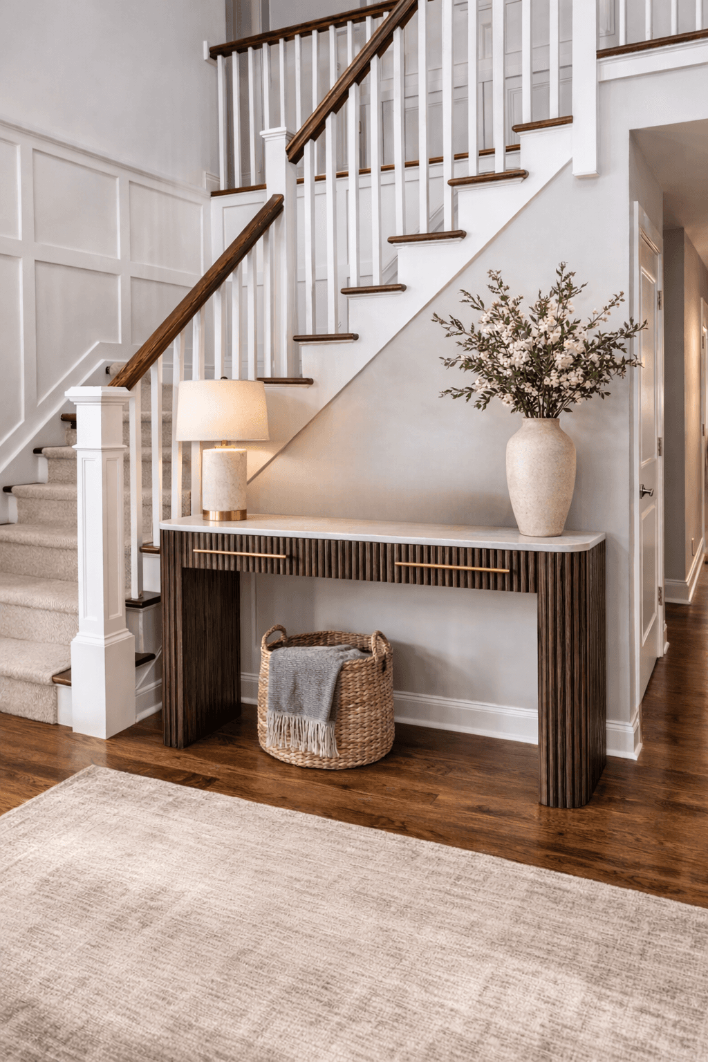 Fluted wood console table with marble top, lamp, and floral vase under a white staircase.