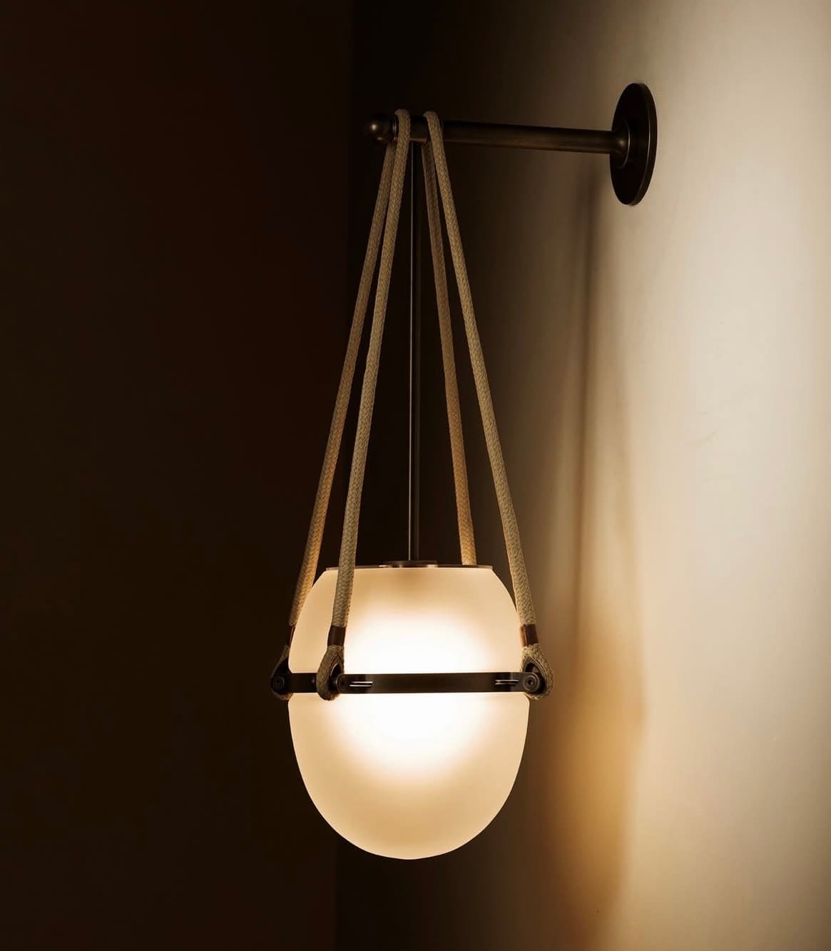 Frosted glass globe sconce suspended by thick ropes from a dark bronze wall arm.